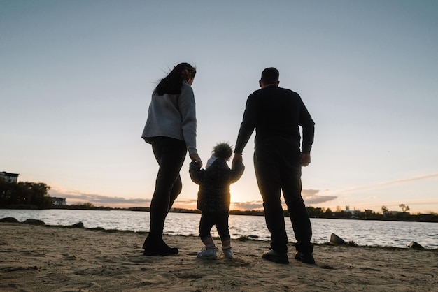 Dziecko w rękach rodziców przytula tatę do mamy i patrzy na słońce Szczęśliwy rodzinny spacer po plaży o zachodzie słońca Córka mama tata spacer po plaży Rodzinne fantazje marzenia z dzieciństwa Widok z tyłu