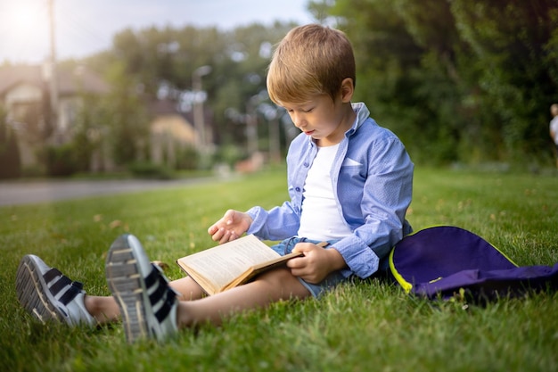 Dziecko w wieku szkolnym siedzi na zielonej trawie w parku z plecakiem i książką czytającą książkę