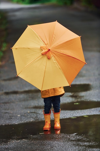 Dziecko w żółtych wodoodpornych butach z płaszczem i parasolem bawiące się na zewnątrz po deszczu