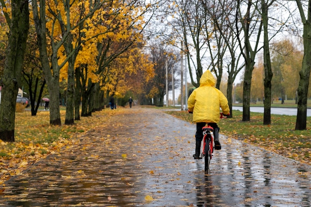 Dziecko w żółtym płaszczu jeździ na rowerze w jesiennym parku podczas deszczu. Widok z tyłu.