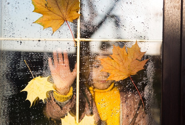 Dziecko wygląda przez okno domu na zewnątrz jesienna pogoda mokra szyba z kroplami po deszczu żółte liście klonu przyklejone do okna jesienny nastrój domowy komfort