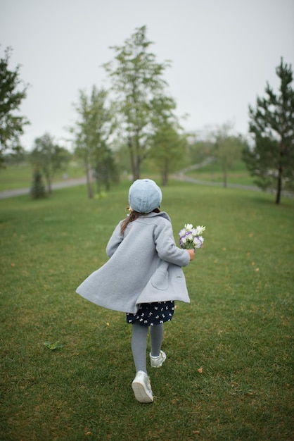 Dziecko z kwiatami w dłoni i szarym płaszczu spaceruje po parku