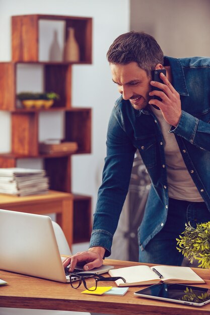 Dzielenie Się Dobrą Wiadomością. Przystojny Młody Mężczyzna Rozmawia Przez Telefon Komórkowy I Patrząc Na Swojego Laptopa Stojąc Przy Biurku W Biurze