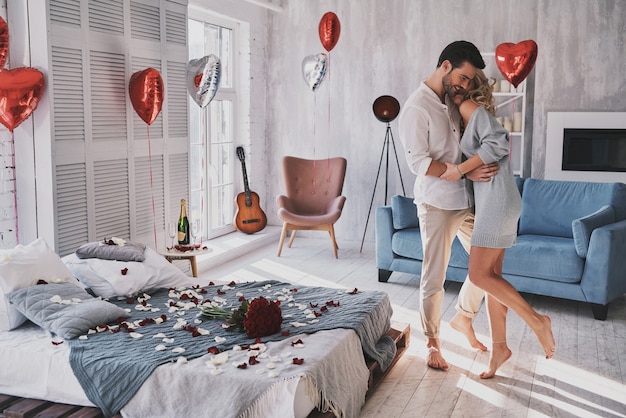 Dzień z okazji ich miłości. Pełna długość pięknej młodej pary obejmującej się i uśmiechającej stojąc w sypialni pełnej balonów