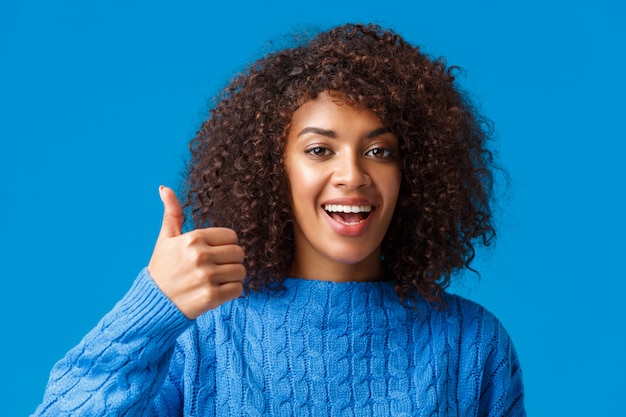 Dziewczyna agresuje z tobą. Close-up portret śliczna afroamerykańska kobieta z afro, kędzierzawa fryzura pokazuje kciuk w zatwierdzeniu, jak lub akceptuje plan, uśmiecha się i kiwa głową w porozumieniu, niebieski