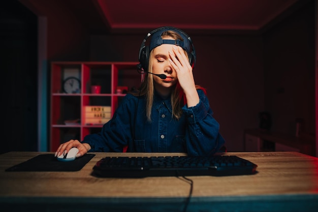 Zdjęcie dziewczyna-gracz siedzi w nocy przy komputerze i pokazuje gest facepalm, spuszcza oczy w dół