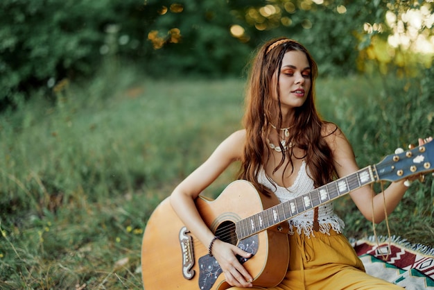 Dziewczyna-hippie grająca na gitarze w ekologicznych ubraniach siedząca na ziemi na zewnątrz w przyrodzie jesienią oglądająca zachód słońca Zdjęcie wysokiej jakości