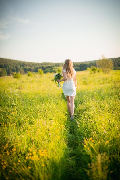 Dziewczyna idzie latem przez pole z kwiatami w białej sukni