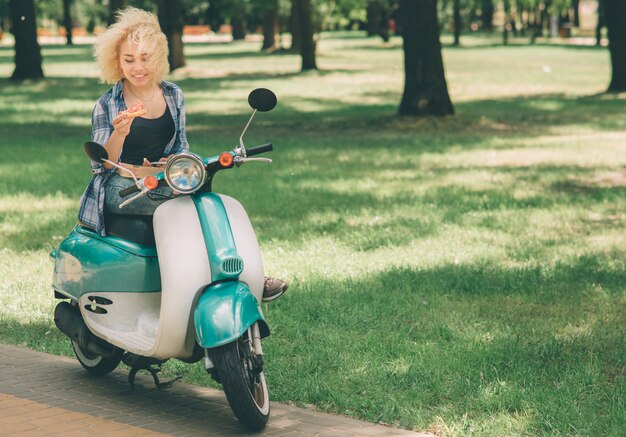 Dziewczyna jedzenie na skuter lub motorower. Dziewczyna jedzenie na skuter lub motorower. Szczęśliwa młoda kobieta trzyma gorącą pizzę w pudełku. Studentka nie ma czasu, będzie jadł w drodze