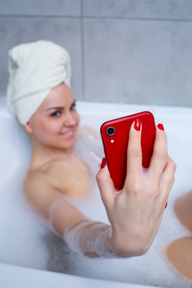 Zdjęcie dziewczyna kobieta z ręcznikiem na głowie leży z lampką szampana w białej wannie i robi sobie selfie przez telefon. w jej rękach dużo pijanego mydła. zrelaksuj się po ciężkim dniu. zabieg relaksacyjny w spa