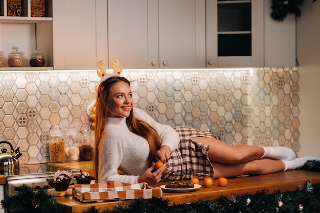 Dziewczyna na Boże Narodzenie leży na kuchennym stole i trzyma w rękach mandarynkę. Kobieta w sylwestra w kuchni leżąca z rogami