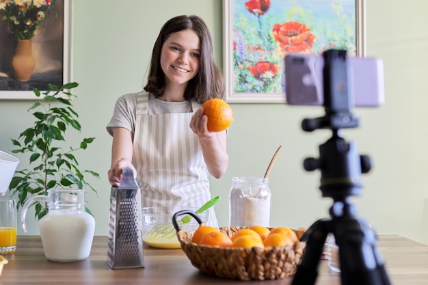 Dziewczyna nastolatek blogger żywności nagrywania przepis na gotowanie pomarańczowych naleśników. Kobieta w domu w kuchni, wyciska sok, uśmiecha się i patrzy na aparat, hobby i wypoczynek, zdrowe smaczne domowe jedzenie koncepcja