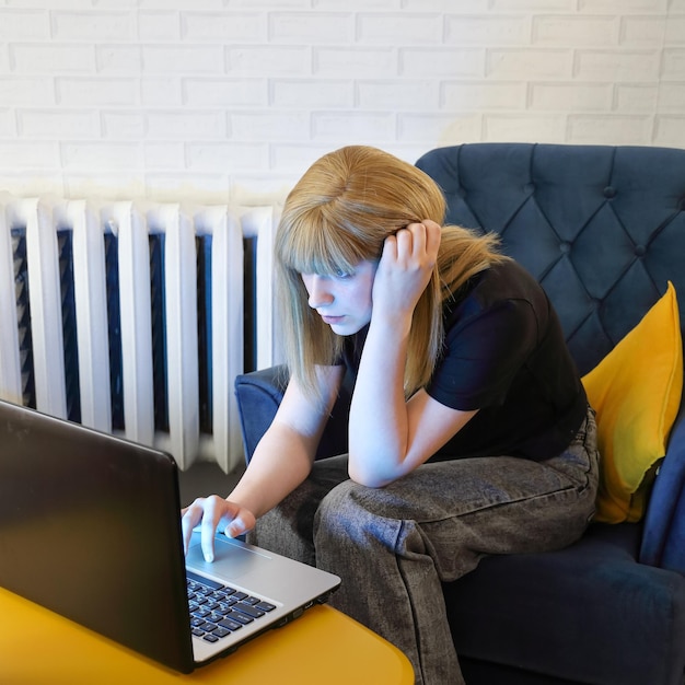 Dziewczyna nastolatka siedzi na krześle przy ścianie za żółtym stołem i robi coś w laptopie