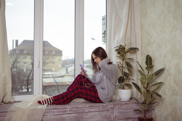 Dziewczyna nastolatka siedzi w oknie z telefonem w ręku i słucha czegoś w słuchawkach. Samotność. Młoda kobieta wygląda przez okno