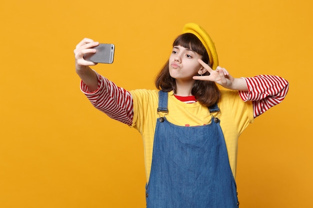 Dziewczyna nastolatka we francuskim berecie, dżinsowa sukienkę robi selfie strzał na telefon komórkowy, pokazując znak zwycięstwa dmuchanie ustami na białym tle na żółtym tle. Koncepcja życia emocje ludzi. Makieta miejsca na kopię.