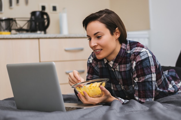 Dziewczyna Ogląda Film Na Komputerze I Kładzie Się Na łóżku, Jedząc Frytki
