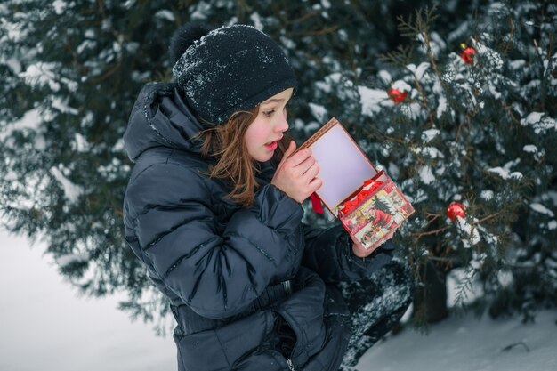 Dziewczyna Otwiera Czerwone Pudełko Z Prezentem. Dziecko Znalazło Prezent Pod Drzewem Na Ulicy. śnieg W Mieście.