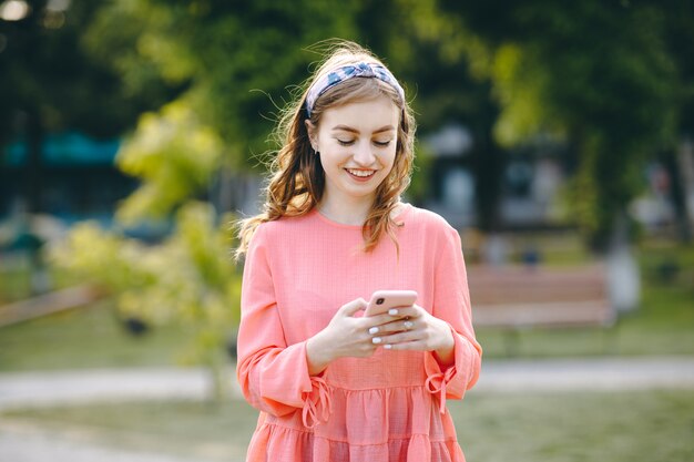 Dziewczyna Się śmieje I Czyta Coś Na Swoim Smartfonie. Rozochocona Młoda Kobieta Komunikuje Na Smartphone Podczas Gdy Chodzący W Parku