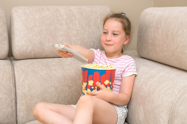 dziewczyna siedzi na kanapie z dużą szklanką popcornu zmienia kanały telewizyjne na pilocie z uśmiechem