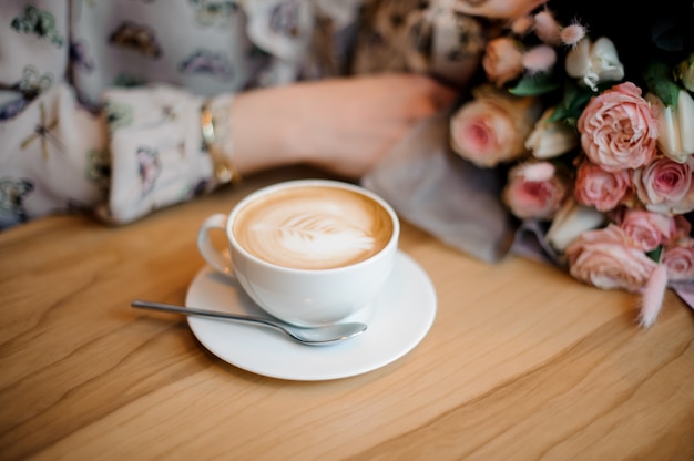 Dziewczyna siedzi przy drewnianym stole z filiżanką kawy i piękny bukiet