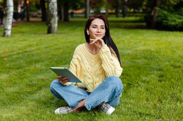 Dziewczyna siedzi w parku i jest zaręczona z iPadem. młoda brunetka siedzi na trawie i zagląda do tabletu. dziewczyna w jesiennym parku.