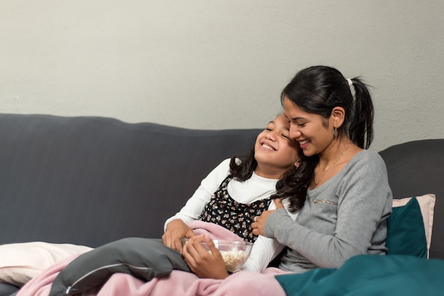 Dziewczyna spoczywa na matce na kanapie, oglądając telewizję i śmiejąc się razem jedząc popcorn. Zdjęcie z miejscem na tekst po lewej stronie.