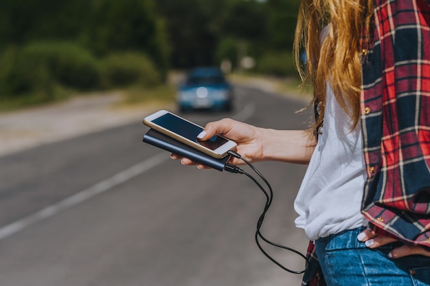 Dziewczyna Trzyma W Dłoni Power Bank Z Telefonem. Na Tle Drogi Z Samochodem I Naturą.