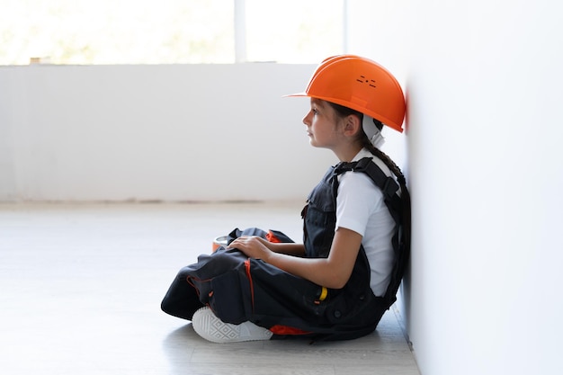Dziewczyna ubrana jest w kombinezon i pomarańczowy hełm ochronny na głowę. siedząc na podłodze, odwracając wzrok.