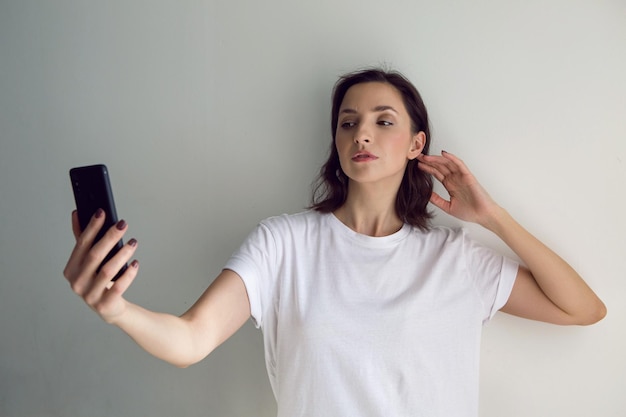 Dziewczyna w białej koszulce robi selfie na czarnym telefonie na białą ścianę domu