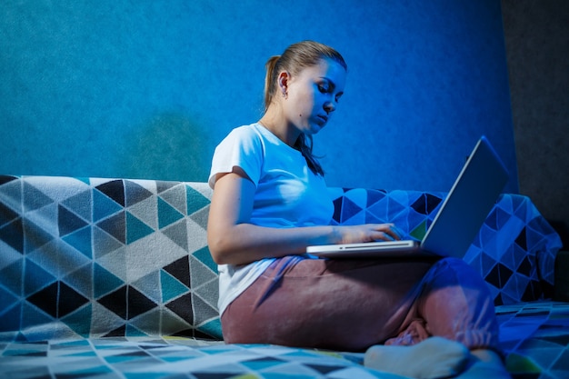 Dziewczyna w białej koszulce siedzi w domu na kanapie z laptopem na kolanach i patrzy w ekran. Czyta nowe ważne informacje. Szukaj wiadomości