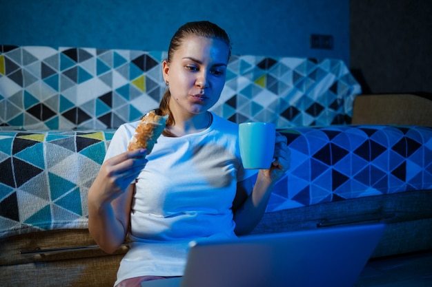 Dziewczyna w białej koszulce siedzi w domu w ciemności na kanapie i ogląda film na swoim laptopie. Zjada rogalika i pije herbatę