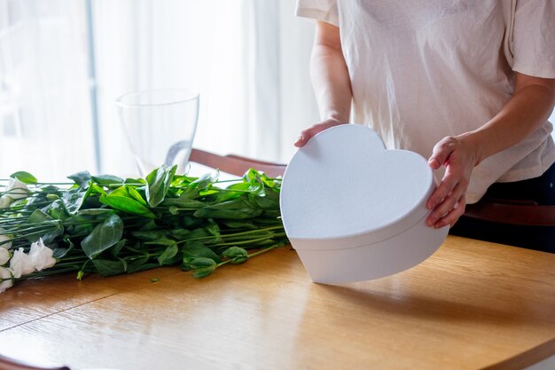 Dziewczyna w białej koszulce trzyma na kuchennym stole białe pudełko w kształcie serca obok bukietu białych róż. Koncepcja stylu życia