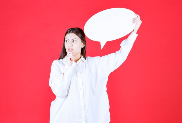Zdjęcie dziewczyna w białej koszuli trzyma owalną tablicę informacyjną i wygląda na zdezorientowaną i zamyśloną
