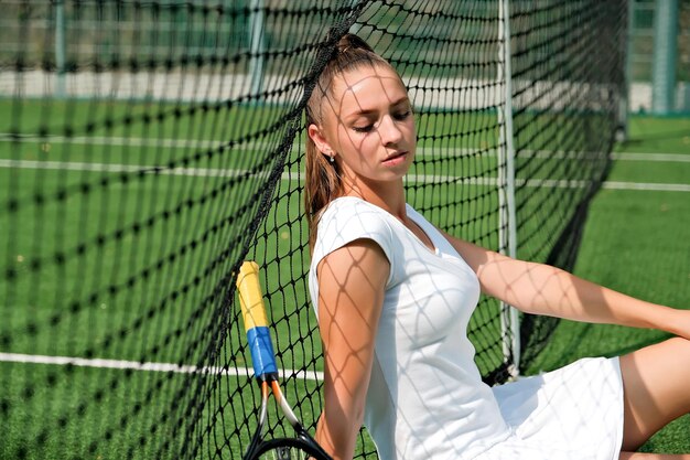 Dziewczyna w białej sportowej sukience siedzi na korcie tenisowym i rakiecie