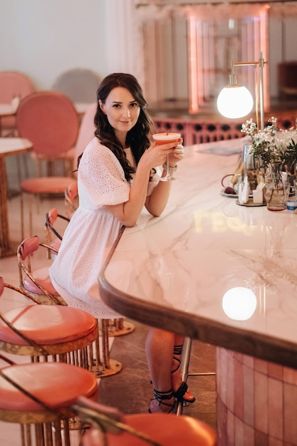 Dziewczyna w białej sukience siedzi przy barze w kawiarni i pije koktajl