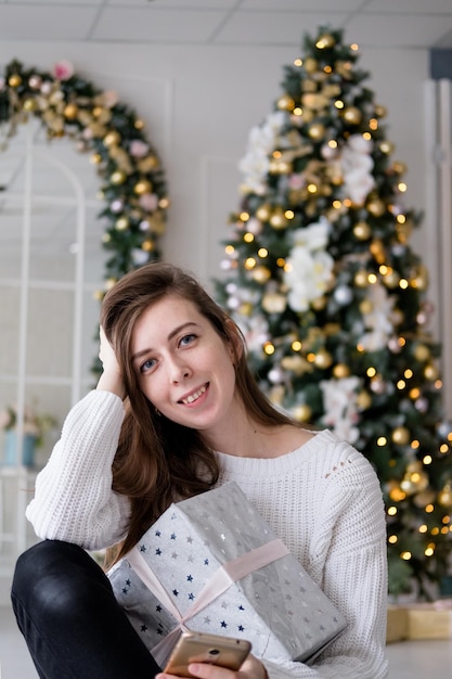 Dziewczyna w białym swetrze siedzi z prezentem na tle choinki. Otrzymanie pudełka z kokardą na Boże Narodzenie