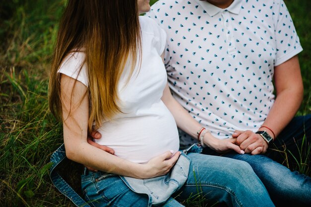 Zdjęcie dziewczyna w ciąży i jej mąż przytulić, trzymać się za ręce, na brzuchu, siedząc na trawie na zewnątrz w powierzchni ogrodu