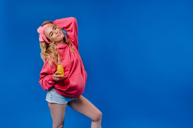 Dziewczyna w ciąży w różowe ubrania z butelką soku na niebieskim tle.