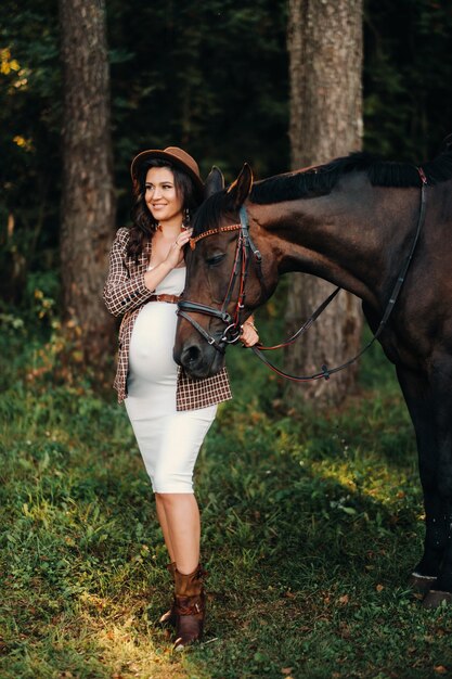 Dziewczyna w ciąży z dużym brzuchem w kapeluszu obok koni w lesie w przyrodzie. Stylowa dziewczyna w białych ubraniach i brązowej kurtce.