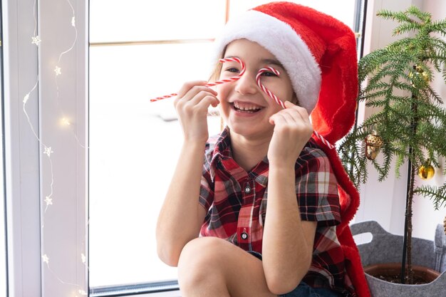 Dziewczyna w czapce Mikołaja siedzi na parapecie domu w pobliżu choinki i przykłada do oczu laskę cukrową jak okulary Dziecko bawi się i robi miny, czekając na Boże Narodzenie i Nowy Rok