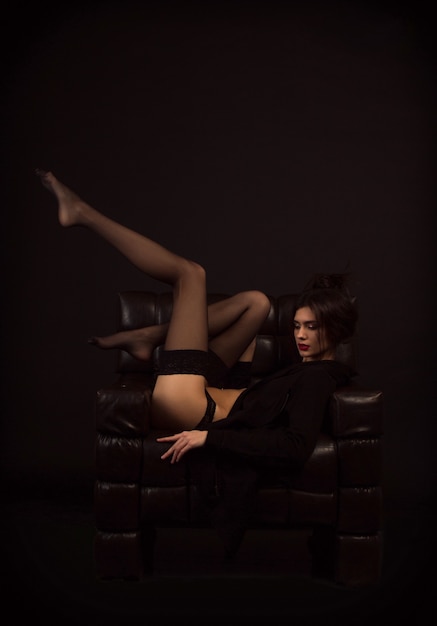 Dziewczyna w czarnych pończochach siedzi w skórzanym fotelu z podniesionymi nogami. seksualnie
