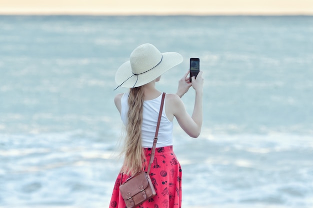 Dziewczyna w czerwieni spódnicie i kapeluszu robi selfie na plaży na tle morza i nieba. Widok z tyłu