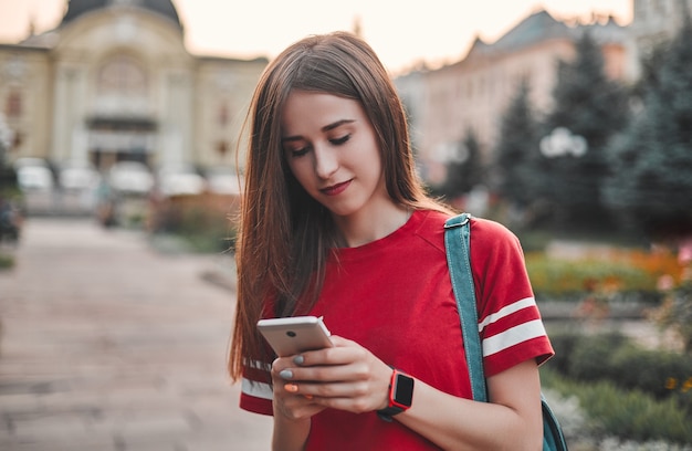 Dziewczyna w czerwonej koszulce brunetka z plecakiem pisze przez telefon na tle miejskiego placu