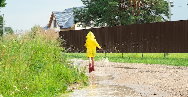Dziewczyna w czerwonych gumowych butach i żółtym płaszczu przeciwdeszczowym biegnie przez kałuże po deszczu we wsi Lato wolność dzieciństwo