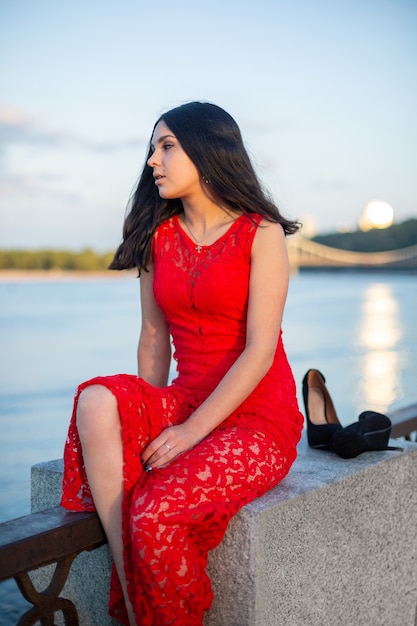 Dziewczyna w długiej czerwonej sukience siedzi na płocie nabrzeża rzeki.
