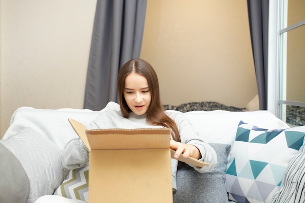Dziewczyna w domu na kanapie z kartonowym pudełkiem otwiera paczkę, zdziwiona