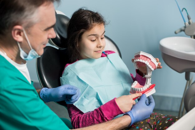 Dziewczyna W Fotelu Dentystycznym Siedzi Ze Swoim Dentystą Pediatrycznym, Pokazując Właściwe Szczotkowanie Zębów, Używając Modelu Szczęki I Szczoteczki Do Zębów W Gabinecie Stomatologicznym