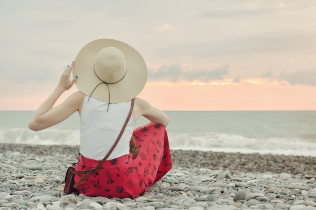 Dziewczyna w kapeluszu siedzi na kamienistej plaży. Widok z tyłu. Czas zachodu słońca