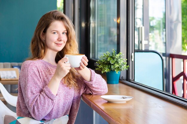 Dziewczyna w kawiarni przy filiżance kawy, uśmiechając się i pijąc latte.