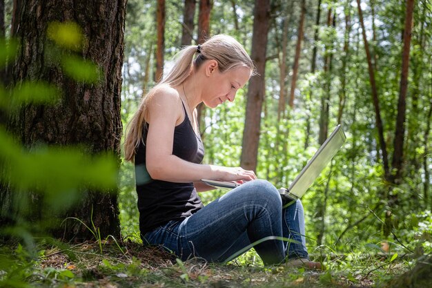 Zdjęcie dziewczyna w lesie siedzi oparta na drzewie i pracuje na laptopie.